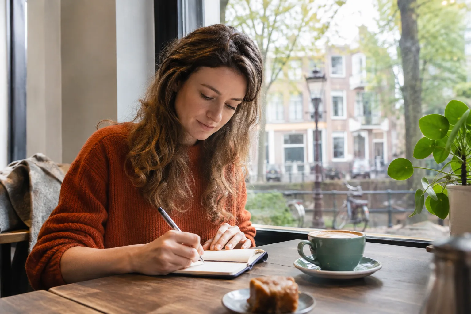 Een hand vult enkele regels in een klein zaknotitieboek in tijdens een koffiepauze bij het raam.