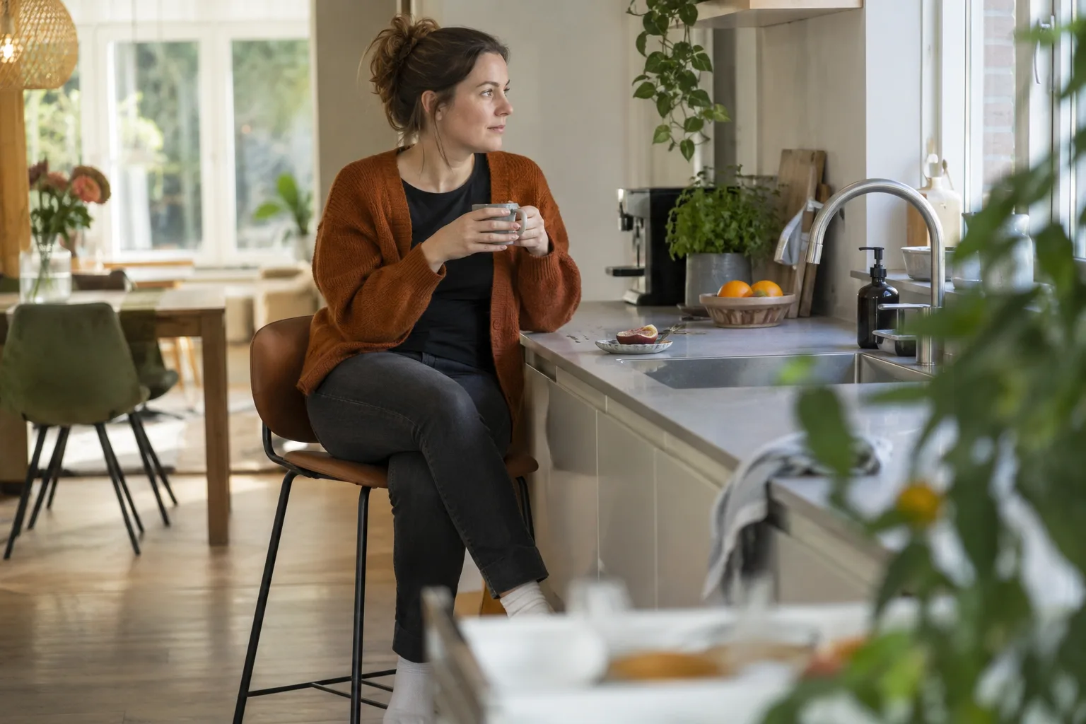 Een vrouw zit tijdens een korte dagelijkse taak rustig op een hoge keukenkruk bij het aanrecht, met ruimte om te pauzeren.