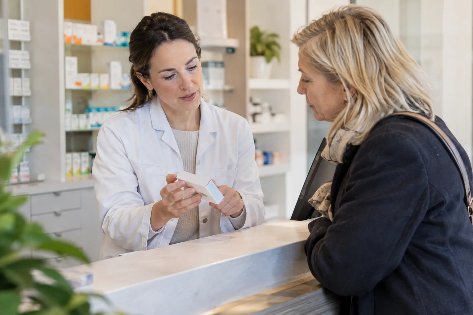 Een apotheker bespreekt een gesloten medicijndoosje met een volwassene aan een rustige balie, tekst onleesbaar.