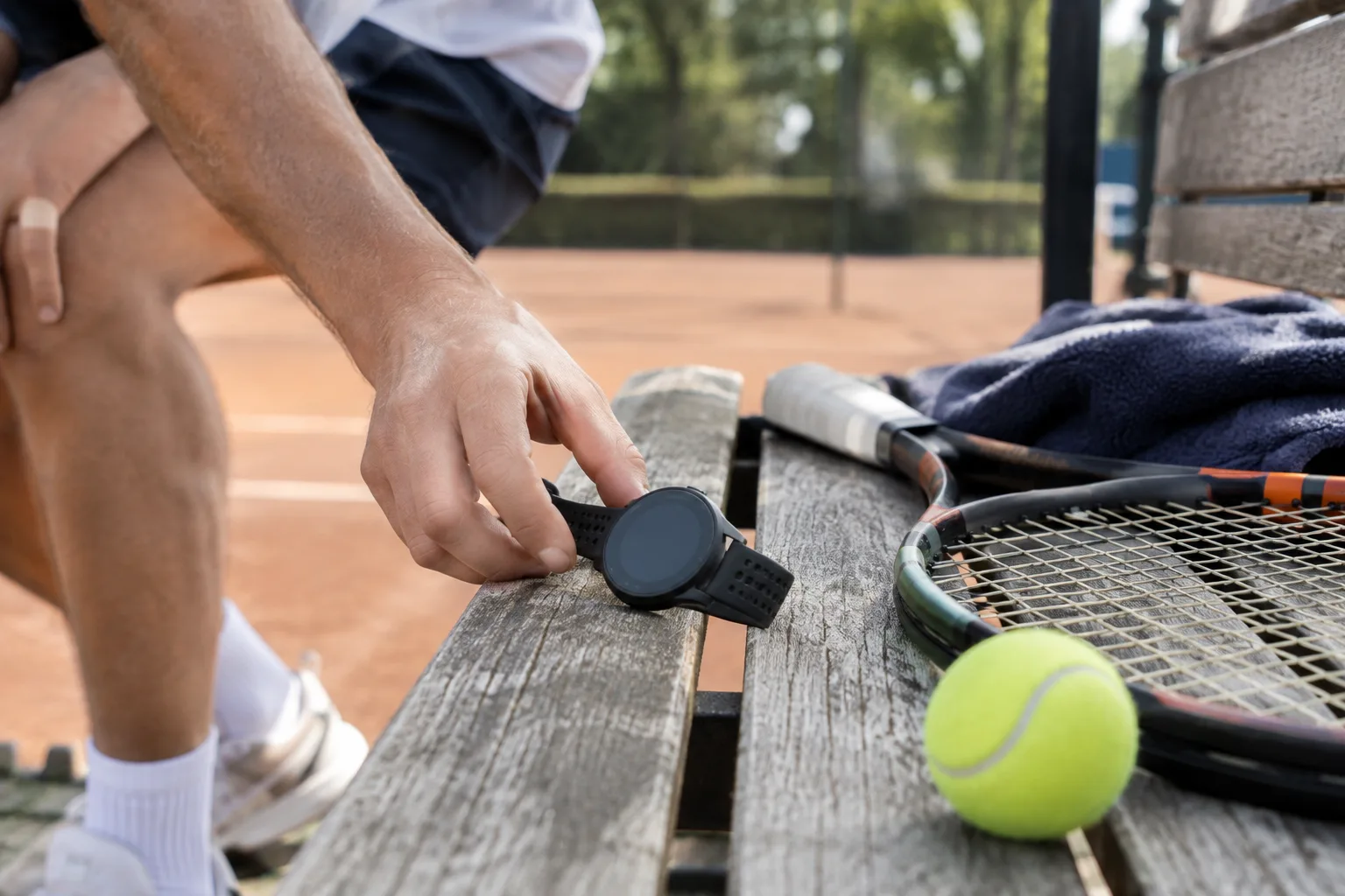Een hand legt een sporthorloge bewust naast een tennisracket op een bankje, in natuurlijk daglicht.