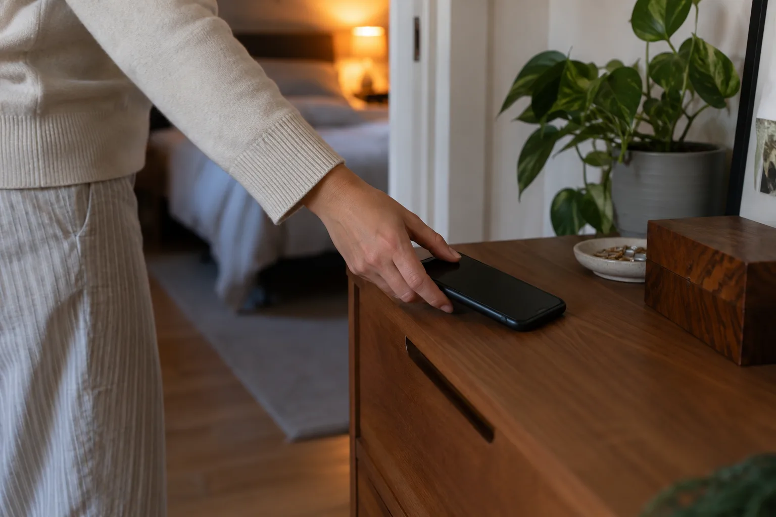 Een hand legt een smartphone met donker scherm buiten bereik op een dressoir, naast een slaapkamerdeur.