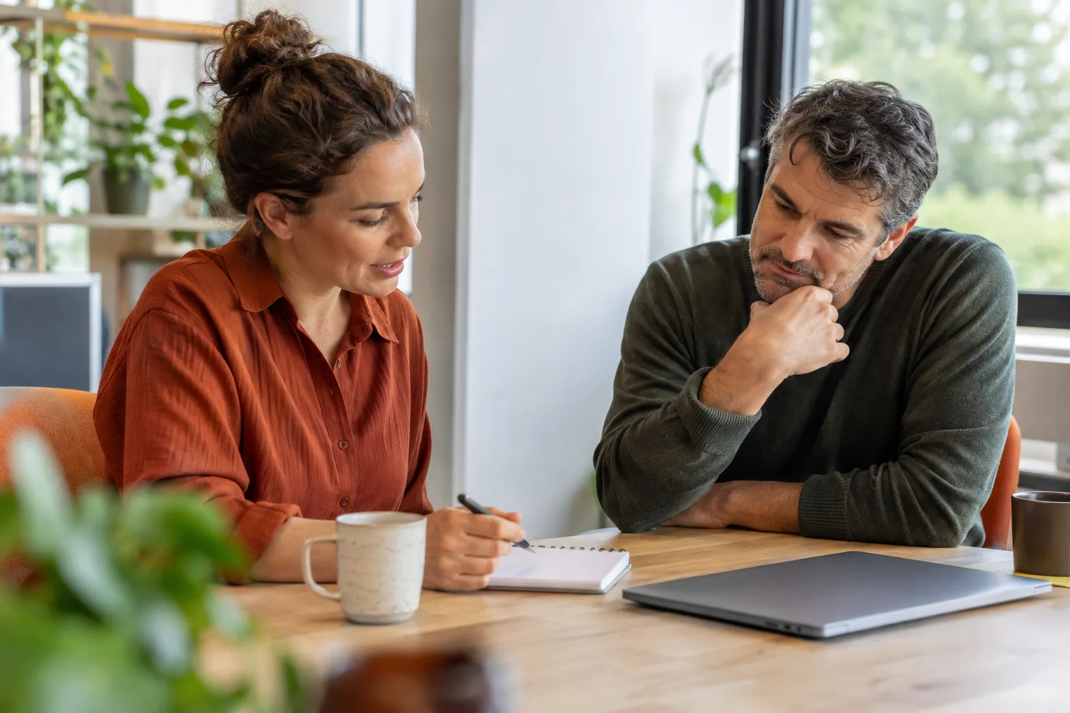 Twee collega's bespreken een kleine takenlijst aan een rustige werktafel met een gesloten laptop.