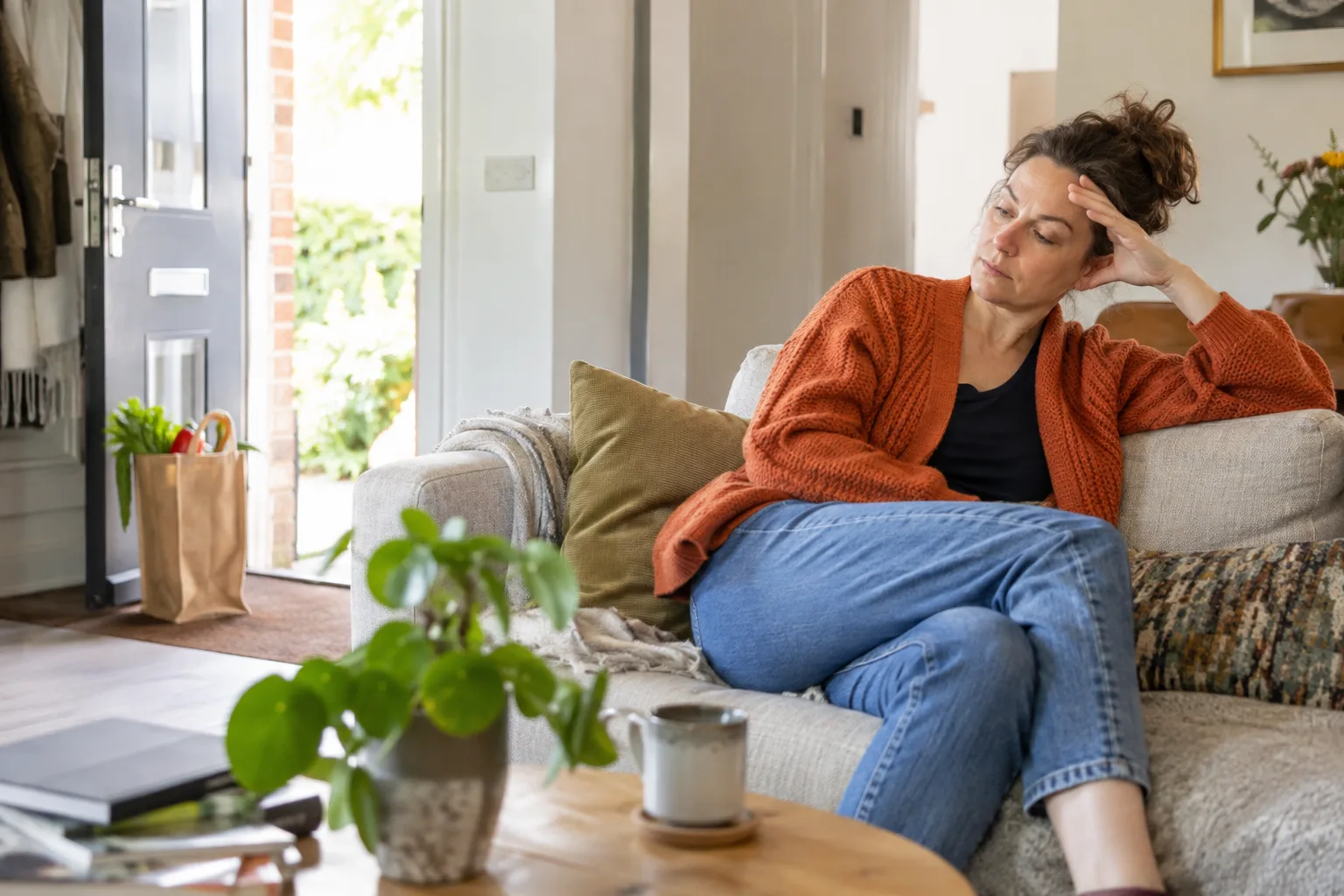 Een volwassen vrouw rust in een lichte woonkamer na een gewone boodschap; een kleine boodschappentas staat bij de deur.