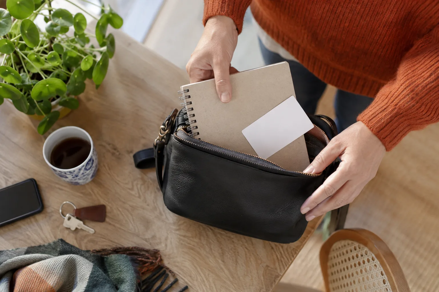 Een hand legt een notitieboek met losse vragen en een afspraakkaartje in een kleine tas, helder bovenaanzicht.